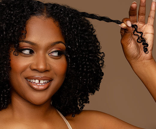Smiling woman with short coily natural hair stretching a defined curl, showcasing healthy, moisturized curls with Design Essentials haircare.