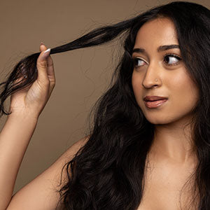 Woman with long dark wavy hair pulling a strand forward, looking to the side against a neutral background.