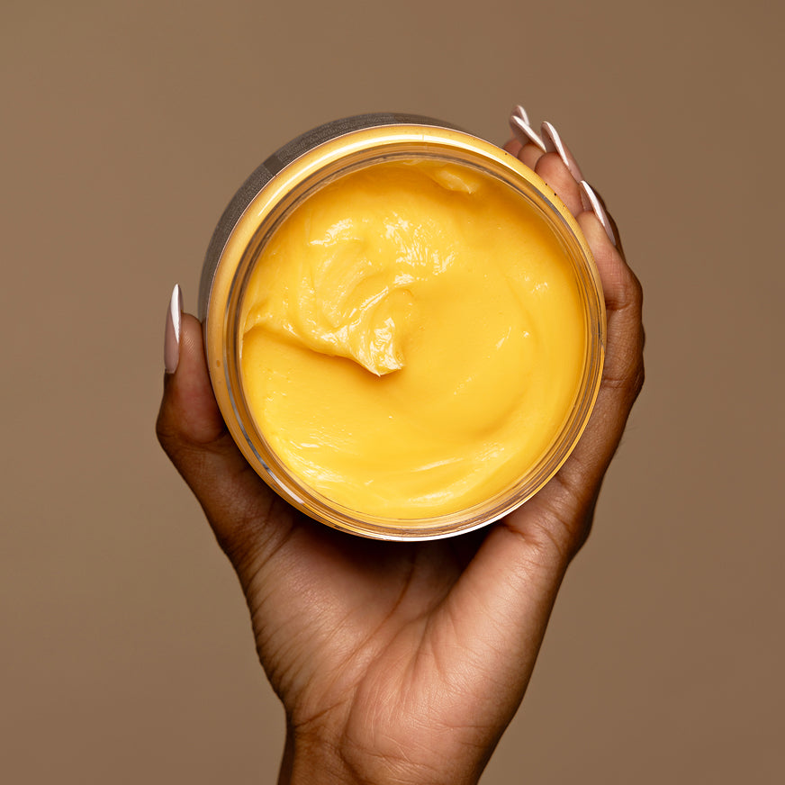 Hand holding up an open jar of Honey Crème Moisture Retention Masque, a rich golden yellow hair cream viewed from above