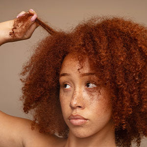 Young woman with red curly hair and freckles gently pulling at a curl while looking off to the side. 
