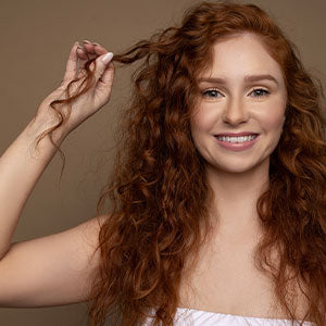 Woman with long red curly hair smiling while holding a curl of her hair in front of a brown background. 